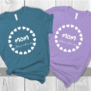 May include: Two t-shirts with a white circle of hearts around the word "MOM" and the text "Happy Mother's Day". The shirt on the left is deep teal and the shirt on the right is lilac.