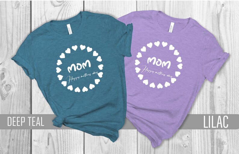 May include: Two t-shirts with a white circle of hearts around the word "MOM" and the text "Happy Mother's Day". The shirt on the left is deep teal and the shirt on the right is lilac.