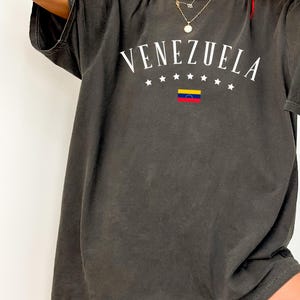 May include: A dark gray oversized t-shirt with a white graphic print of the word "VENEZUELA" in an arc with five white stars on each side of the word and a small Venezuelan flag in the center.