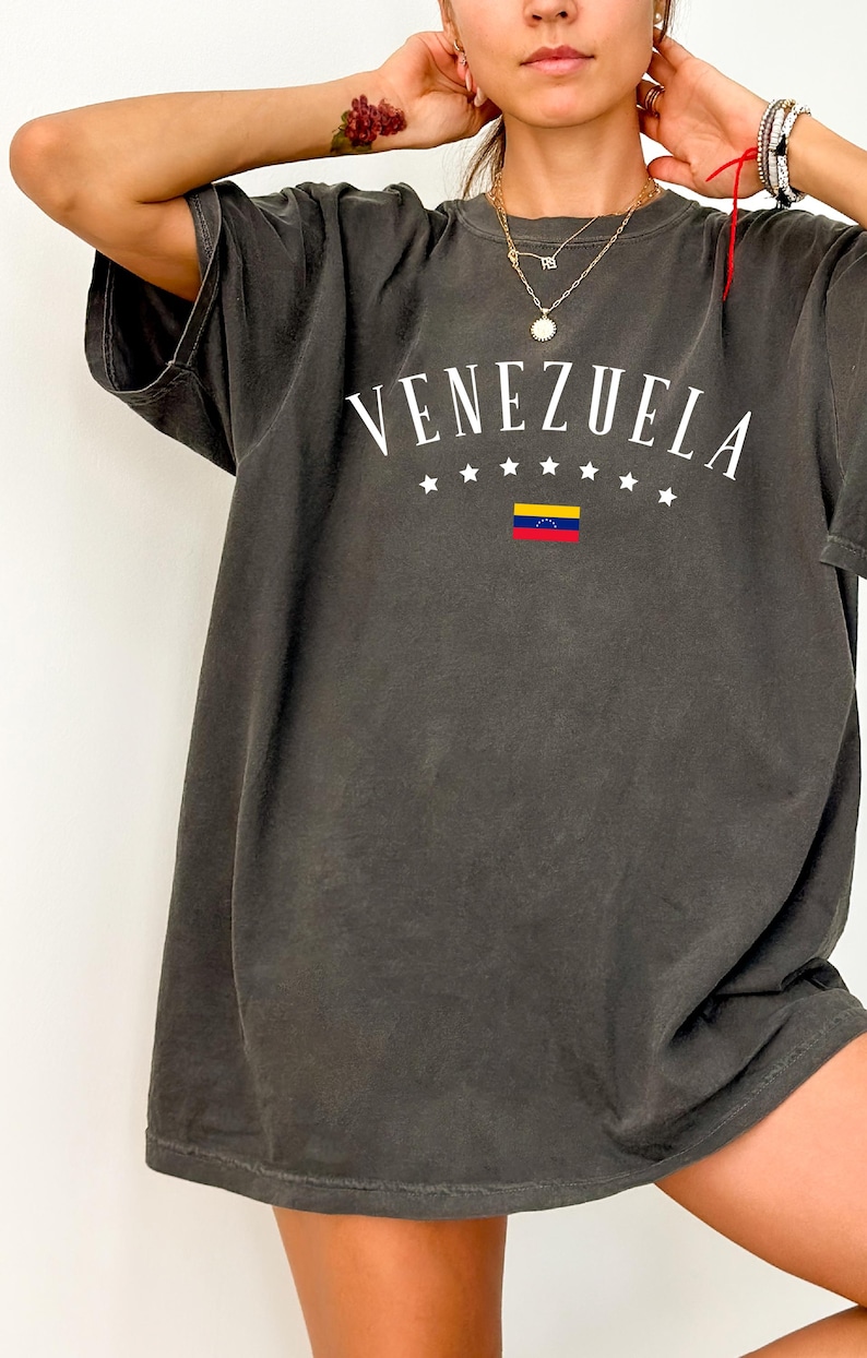 May include: A dark gray oversized t-shirt with a white graphic print of the word "VENEZUELA" in an arc with five white stars on each side of the word and a small Venezuelan flag in the center.