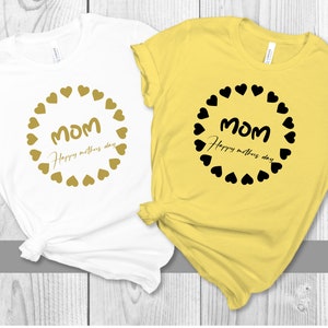 May include: Two t-shirts with the text "Mom Happy Mother's Day" in a circle of hearts. One shirt is white with gold hearts and text, the other is yellow with black hearts and text.