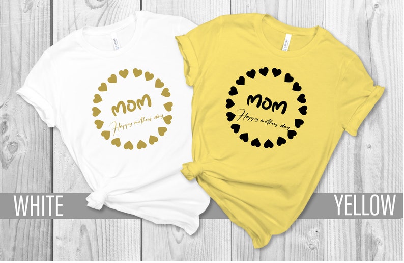 May include: Two t-shirts with the text "Mom Happy Mother's Day" in a circle of hearts. One shirt is white with gold hearts and text, the other is yellow with black hearts and text.