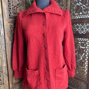 May include: A red button-up cardigan with long sleeves and two patch pockets. The cardigan is made of a soft, textured fabric.