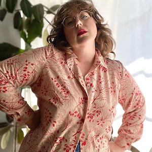 May include: A beige button-up shirt with a red leaf pattern. The shirt has long sleeves and a collared neckline.
