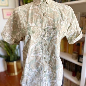 May include: A short-sleeved button-up shirt with a green and white abstract print. The shirt has two flap pockets on the chest and a collar.
