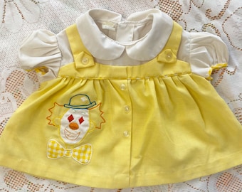 Pode incluir: Um vestido amarelo para bebê com gola branca e um palhaço bordado na frente. O vestido tem fecho de botões e é feito de um tecido macio e confortável.