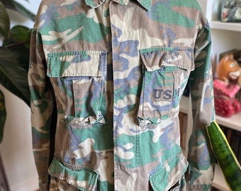 Puede incluir: Una chaqueta de camuflaje con un parche USMC en el bolsillo. La chaqueta tiene varios bolsillos y un cierre de botones en la parte delantera.