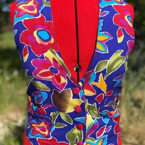 May include: A sleeveless vest with a vibrant floral pattern in shades of red, pink, yellow, blue, and green on a deep blue background. The vest has a V-neck and button closure.