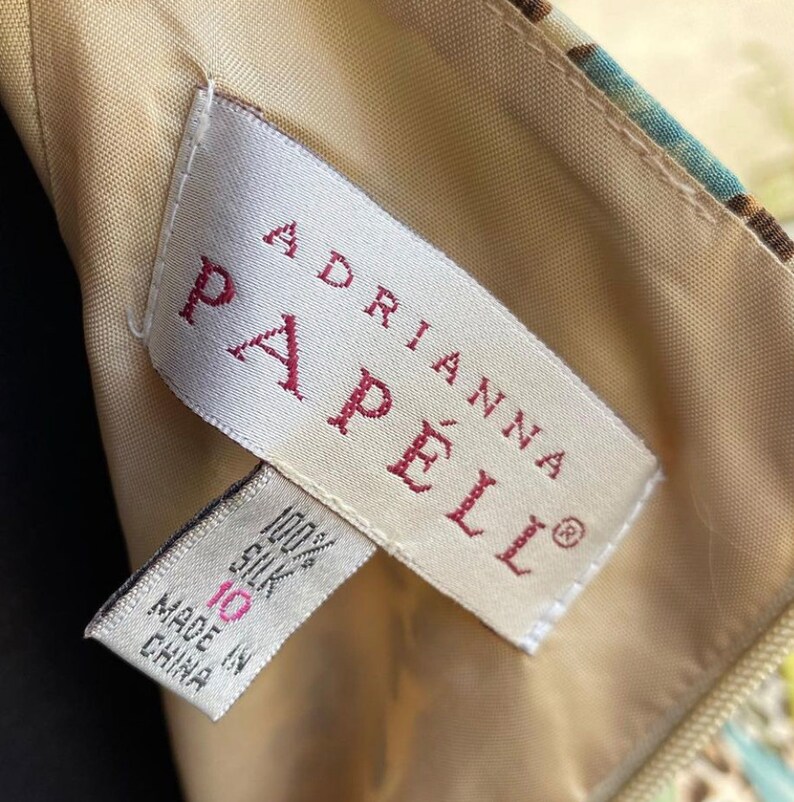 Puede incluir: Una etiqueta blanca con texto rojo que dice "Adrianna Papell" y "100% Seda" con una etiqueta de talla 10 y el texto "Hecho en China".
