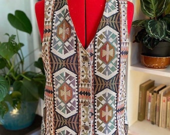 May include: A brown, beige, and green patterned vest with a V-neck and button closure. The vest is made of a woven fabric with a geometric design.