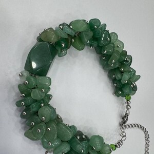 Pode incluir: Uma pulseira com numerosas pedras verdes de aventurina polida e uma conta de pedra verde maior, lisa e retangular. As pedras são intercaladas com pequenas contas de metal prateado e ligadas a uma corrente de metal prateado.