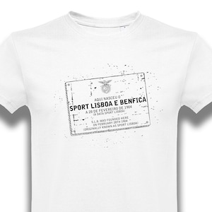 May include: White t-shirt featuring a black and white graphic print of a vintage stamp that reads "AQUI NASCEU O SPORT LISBOA E BENFICA A 28 DE FEVEREIRO DE 1904 (A DATA SPORT LISBOA) S.L.B. WAS FOUNDED HERE ON FEBRUARY 28TH 1904 (ORIGINALLY KNOWN AS SPORT LISBOA)"