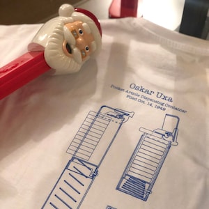 Puede incluir: Una camiseta blanca con un diseño azul y blanco de un contenedor de dispensación de artículos de bolsillo. El diseño incluye el texto "Oskar Uxa", "Pocket Article Dispensing Container", "Filed Oct. 14, 1949", "Patented Dec. 2, 1952", y "N°. 2.620.061".