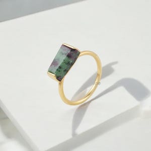Puede incluir: Un anillo dorado con una piedra rectangular con tonos verdes y granates. La piedra está engastada en un bisel dorado, y la banda del anillo es de oro pulido. El anillo se muestra sobre una superficie blanca.
