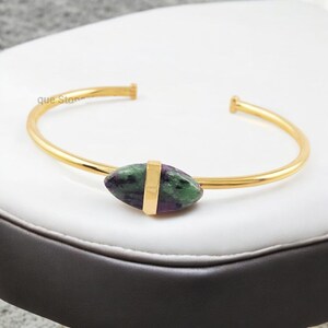 Pode incluir: Pulseira aberta dourada com uma pedra central ovalada em tons de verde e roxo. A pedra é fixada por uma faixa dourada. A pulseira é exibida em uma superfície branca.