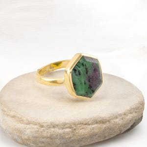 Puede incluir: Un anillo de oro con una gran gema hexagonal. La gema presenta una mezcla de colores verdes y granates. El anillo está sobre una superficie clara y texturizada. La banda y el engaste del anillo son de color dorado brillante.