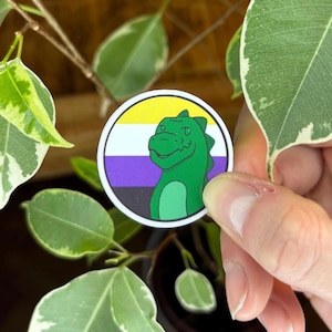 May include: A circular sticker featuring a non-binary pride flag with horizontal yellow, white, purple, and black stripes. A green cartoon dinosaur with a light green belly is positioned in the foreground, facing slightly to the left.