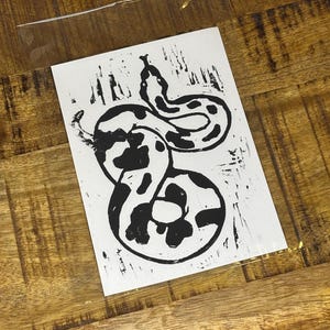 May include: A rectangular white card featuring a black ink block print of a coiled snake with a spotted pattern. The snake is depicted in a stylised manner with visible texture from the printing process, enclosed in a clear plastic sleeve.