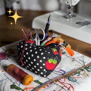 May include: A black polka dot and strawberry print sewing caddy filled with sewing tools. The caddy holds scissors, a pen, needles, and spools of thread. A spool of orange thread sits in front of the caddy. A sewing machine is in the background.