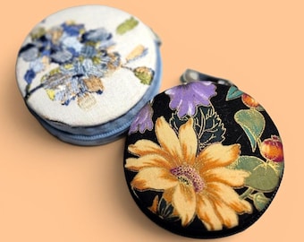 May include: Two round, zippered fabric cases. One case features a white background with blue and green floral embroidery. The second case has a black background with a large yellow sunflower, purple petals, and green leaves printed on the fabric.