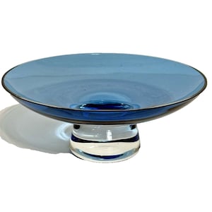 May include: A blue glass pedestal bowl with a round, shallow dish and a clear and blue striped base. The bowl has a dark blue rim and is set against a white background. The bowl is approximately 10 inches in diameter.