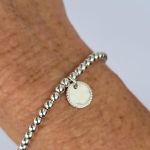 Könnte beinhalten: Silbernes Perlenarmband mit einem runden Anhänger. Der Anhänger hat einen strukturierten Rand und eine glatte Mitte. Das Armband besteht aus kleinen, runden Perlen und wird am Handgelenk getragen.
