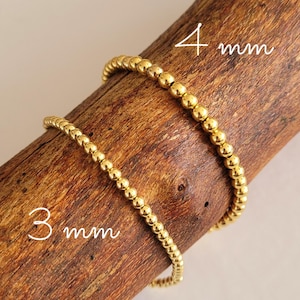 Könnte beinhalten: Zwei goldene Perlenarmbänder auf einer Holzoberfläche. Das obere Armband hat 4 mm Perlen und das untere Armband hat 3 mm Perlen.