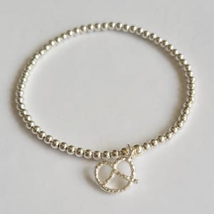 Könnte beinhalten: Ein silbernes Armband mit kleinen, runden Perlen und einem herzförmigen Brezel-Anhänger. Der Anhänger ist detailliert gestaltet und verleiht dem Schmuckstück eine besondere Note. Ein zartes und elegantes Accessoire.