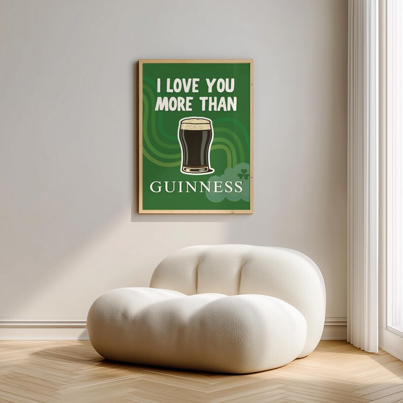 May include: A framed green poster with the text "I LOVE YOU MORE THAN GUINNESS" and an illustration of a pint of Guinness. The poster is hanging on a light grey wall above a white, rounded, upholstered chair.