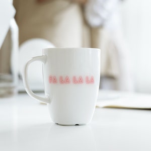 Könnte beinhalten: Weiße Keramiktasse mit der Aufschrift "Fa La La La" in pinkfarbenem Neonlicht.