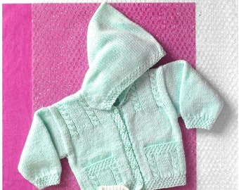 May include: Two knitted baby sweaters, one in light blue and one in white, with a hood on the blue sweater. The sweaters are laid out on a pink bubble wrap background. The text "UKHKA 67" is in the top right corner of the image. The text "UKHKA" is in the bottom left corner of the image. The text "www.ukhanking.com" is in the bottom right corner of the image. The text "UK HAND KNITTING ASSOCIATION" is below the website address.