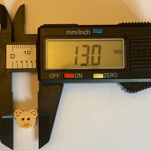 Pode incluir: Um bot&atilde;o de madeira em forma de urso de cor castanho, com um di&acirc;metro de 13,0 mm, medido com um paqu&iacute;metro digital.