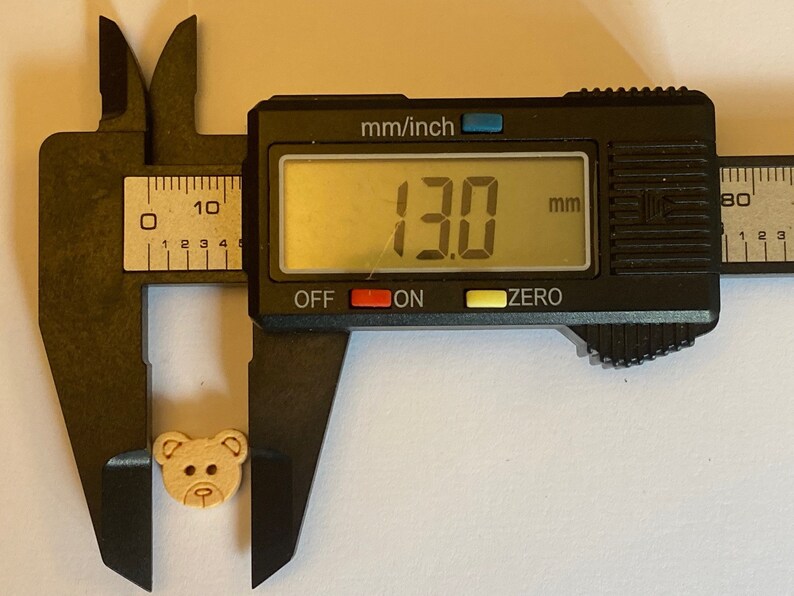 Pode incluir: Um bot&atilde;o de madeira em forma de urso de cor castanho, com um di&acirc;metro de 13,0 mm, medido com um paqu&iacute;metro digital.
