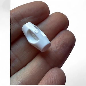 May include: A small, white plastic cord lock with a cylindrical base and a curved top. The cord lock has a hole for the cord to pass through and a small button to secure the cord. The object is held in a hand.