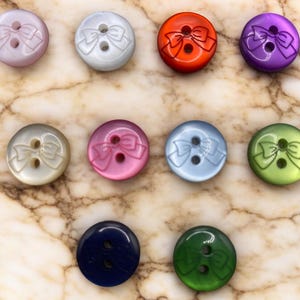 May include: Assortment of 10 colourful buttons with a bow design. The buttons are in various colours including pink, white, red, purple, light blue, green, dark blue, and light green.