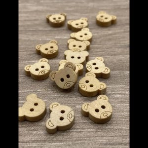 May include: A selection of small, light brown wooden buttons, each crafted in the shape of a teddy bear's face. The buttons feature eye holes, a nose, and ear details. They are scattered on a light brown surface.