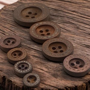 May include: Assortment of brown wooden buttons in various sizes and intricate designs. The buttons have a rustic look and are arranged on a wooden surface.