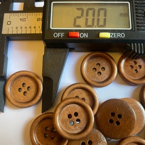 May include: A close-up of a group of 20 mm brown wooden buttons. The buttons have four holes each and are arranged in a random pattern. A caliper is measuring one of the buttons.