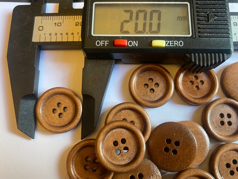 May include: A close-up of a group of 20 mm brown wooden buttons. The buttons have four holes each and are arranged in a random pattern. A caliper is measuring one of the buttons.