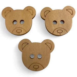 May include: Three light brown wooden buttons shaped like bear faces. Each button features a detailed outline of a bear's face, including ears, eyes, and a nose. The buttons have two holes for sewing.