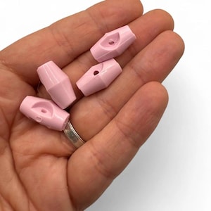May include: Five pale pink toggle clasps. Each clasp is a small, barrel-shaped piece with a hole on one end and a curved slot on the other. The clasps are smooth and have a matte finish. They are held in a hand.