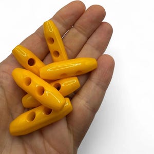 May include: A selection of bright yellow toggle buttons, each with two or three holes. The buttons are oval-shaped and have a glossy finish. They are held in a hand against a white background.