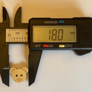 Pode incluir: Um close-up de um bot&atilde;o de madeira em forma de urso medindo 18,0 mm de di&acirc;metro. O bot&atilde;o &eacute; marrom claro com um rosto pintado e olhos pretos.