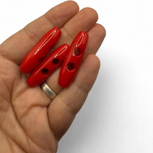 May include: Three bright red toggle buttons with two holes each. The buttons are oval-shaped and have a glossy finish. They are shown against a neutral background.
