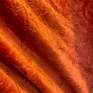 May include: Close-up of a rust-colored fabric with a raised floral pattern. The fabric is folded and has a soft, velvety texture.