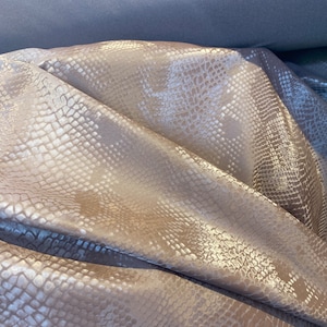 May include: Close-up of a light brown fabric with a snakeskin pattern. The fabric has a subtle sheen and a textured surface.