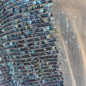 May include: A close-up of a light blue fabric with a pattern of sequins. The sequins are arranged in rows and are iridescent, reflecting different colors of light.