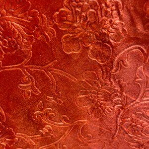 May include: Close-up of a rust-colored fabric with an embossed floral pattern. The fabric has a soft, velvety texture.