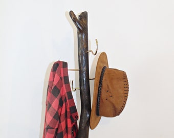 May include: A wooden coat rack with a natural branch design. It has three metal hooks and is decorated with a red and black plaid shirt and a brown cowboy hat.
