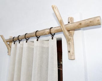 Puede incluir: Una barra de cortina rústica hecha de madera flotante natural. La barra está soportada por soportes de madera y presenta anillos metálicos que sostienen cortinas de lino color crema. El diseño evoca una estética costera o bohemia.